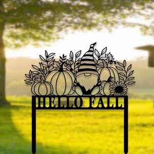 Könnte beinhalten: Schwarzer Metall-Gartenpfahl mit einem Zwerg und Kürbissen, Blättern und einer Sonnenblume. Der Pfahl trägt die Aufschrift "Hello Fall" in schwarzen Buchstaben.
