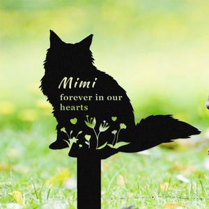 Könnte beinhalten: Schwarze Metallsilhouette einer Katze mit dem Text "Mimi für immer in unseren Herzen" und kleinen Herzen und Blumen. Die Silhouette ist an einem schwarzen Metallpfahl befestigt.