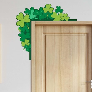 Könnte beinhalten: Eine grüne Kleeblatt-Eckdekoration mit verschiedenen Grüntönen, platziert an einer weißen Wand neben einer Holztür. Die Kleeblätter bilden einen dekorativen Rahmen, ideal für St. Patrick's Day.