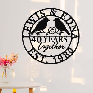 Könnte beinhalten: Schwarzes Metall-Wandbild mit zwei Vogel-Silhouetten auf einem Ast in einem Kreis. Der Text lautet "LEWIS & EDNA", "40 YEARS Together" und "EST. 1980". Ein dekoratives Element für das Zuhause, ideal als Geschenk.