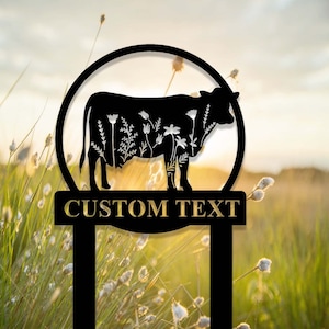 May include: Black metal silhouette of a cow with floral designs, mounted on a black metal stake with a gold plaque that reads "CUSTOM TEXT". The stake is in a field of long grass.