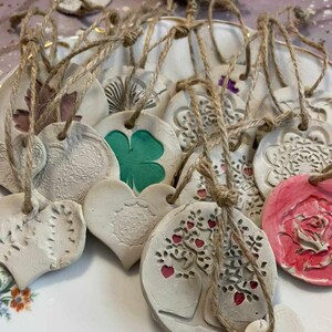 Op de afbeelding: Een verzameling handgemaakte klei-versieringen in verschillende vormen en ontwerpen, waaronder harten, cirkels en klavertjesvier. De versieringen zijn geschilderd in tinten groen, roze en wit, en hebben ingewikkelde details. Elke versiering is bevestigd aan een touwtje van touw.