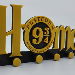 Könnte beinhalten: Ein schwarzer und goldener Schlüsselhalter mit den Worten "Home" und einem runden Emblem mit der Aufschrift "Platform 9 3/4 Kings Cross London".