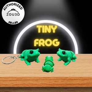 Puede incluir: Tres llaveros de rana verde sobre una superficie de madera. Las ranas son pequeñas y tienen ojos negros. El texto "TINY FROG" se muestra en amarillo sobre las ranas.