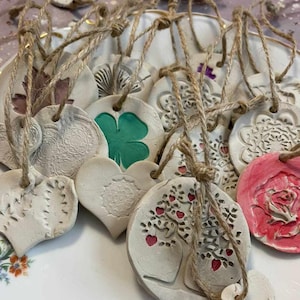 Op de afbeelding: Een verzameling handgemaakte klei-versieringen met verschillende ontwerpen, waaronder harten, bloemen en een boom. De versieringen zijn aan touw geregen en klaar om opgehangen te worden.