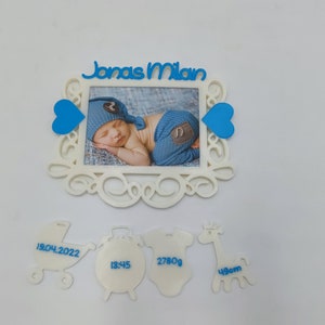 Può includere: Cornice per foto bianca con un cuore blu su ogni lato, una foto di un bambino all'interno e il nome "Jonas Milan" in alto. Sotto la cornice ci sono quattro figure bianche con testo blu: un passeggino con "19.04.2022", un orologio con "13:45", un body con "2780g" e una giraffa con "49cm".