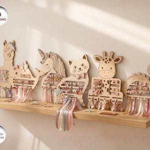 Può includere: Organizzatori di accessori per capelli in legno in sei modelli: lama, unicorno, gatto, giraffa e dinosauro. Ogni organizzatore ha fessure per fermagli per capelli e nastri. Gli organizzatori sono esposti su una mensola di legno contro un muro bianco.