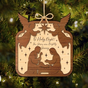 May include: Wooden Christmas ornament with a layered design. The ornament features a nativity scene with angels, stars, and the text "O Holy Night! The Stars are Brightly Shining". It is suspended by a natural twine.