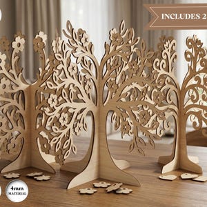 May include: Three light brown wooden tree sculptures with intricate laser-cut branches, leaves, and flower details. The set includes 3 designs and 2 sizes, with 3mm and 4mm material thickness options. Small wooden flower and heart shapes are scattered.