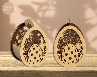 Könnte beinhalten: Zwei hölzerne Ostereier-förmige Boxen mit filigranen Ausschnitten. Eine Box hat einen Griff und die andere ist ein Kerzenhalter. Beide Boxen zeigen Blumenmuster und ein kleineres Ei im Inneren.
