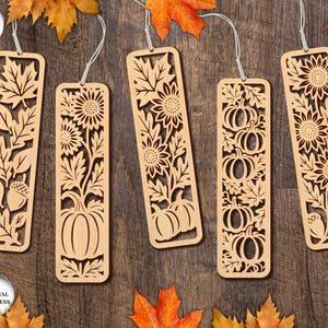 Könnte beinhalten: Fünf Lesezeichen aus Holz mit aufwendigen Ausschnitten. Jedes Lesezeichen zeigt ein anderes herbstliches Design, darunter Blätter, Sonnenblumen, Kürbisse und Eicheln. Die Lesezeichen sind hellbraun und haben eine Schnur zum Aufhängen.