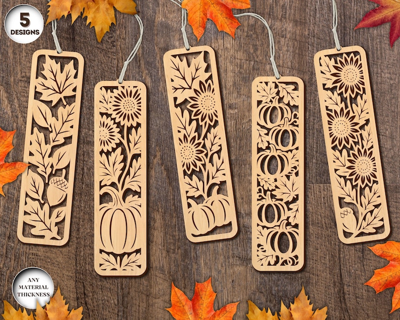 May include: Five wooden bookmarks with intricate cut-out designs. Each bookmark features a different fall-themed design, including leaves, sunflowers, pumpkins, and acorns. The bookmarks are light brown and have a string for hanging.
