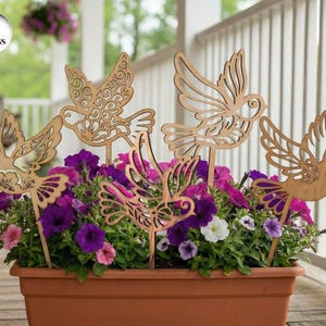 Puede incluir: Estacas de jardín de pájaros de madera en varios diseños se exhiben en una jardinera de terracota llena de petunias moradas, rosas y blancas. Las estacas tienen intrincados detalles recortados. La imagen incluye un gráfico circular que dice "5 DESIGNS".