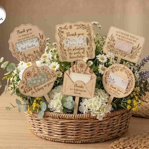 May include: A wicker basket overflowing with white and yellow flowers, adorned with six wooden signs. The signs feature floral designs and phrases like "Thank you for helping me grow" and "Best Teacher."