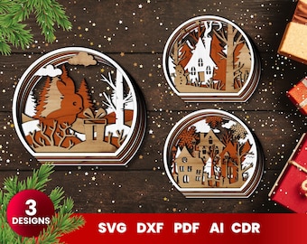 Può includere: Tre ornamenti natalizi in legno a più strati con intricati ritagli. Il primo ornamento presenta un coniglio che tiene un regalo in una scena di foresta innevata. Il secondo ornamento presenta una piccola casa con un camino in una scena di foresta innevata. Il terzo ornamento presenta una casa a due piani in una scena di foresta innevata.