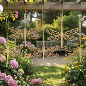 Puede incluir: Seis adornos de madera con forma de libélula con intrincados diseños calados, colgados de cuerdas. Las libélulas son de color marrón claro y están suspendidas sobre un jardín con flores y vegetación.