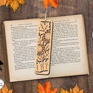 May include: A wooden bookmark with a laser-cut design featuring sunflowers and pumpkins. The bookmark is rectangular and has a string attached. The bookmark is lying on an open book with a cup of coffee nearby.