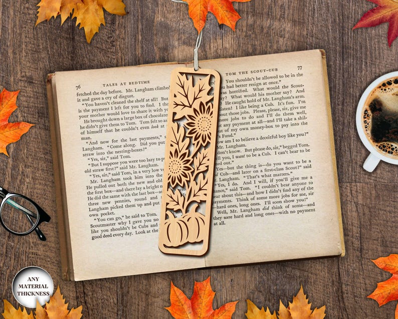 May include: A wooden bookmark with a laser-cut design featuring sunflowers and pumpkins. The bookmark is rectangular and has a string attached. The bookmark is lying on an open book with a cup of coffee nearby.