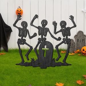 May include: A Halloween garden decoration featuring three black skeleton figures dancing around a tombstone. A black ghost, carved pumpkins, and a spider web complete the spooky scene. The decorations are set on green grass with a white fence backdrop.