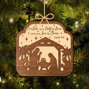 May include: Wooden Christmas ornament with a nativity scene cut-out design. The ornament features a layered design with a light-colored interior and a darker wood frame. Text includes "For Unto us a Child is Born" and "Isaiah 9:6".
