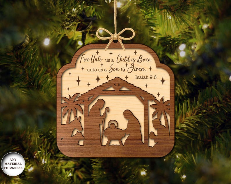 May include: Wooden Christmas ornament with a nativity scene cut-out design. The ornament features a layered design with a light-colored interior and a darker wood frame. Text includes "For Unto us a Child is Born" and "Isaiah 9:6".