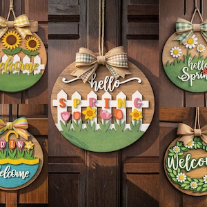 Op de afbeelding: Vijf houten welkomstborden met lentethema. Elk rond bord heeft een ander ontwerp, waaronder zonnebloemen, madeliefjes en bloemstukken, met de woorden "Welcome" of "Hello Spring". Elk bord heeft een decoratieve strik.