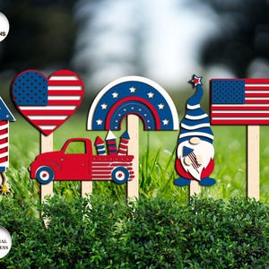 May include: A collection of six patriotic wooden lawn decorations. Designs include a rocket, heart, rainbow, red truck, gnome, and American flag, all featuring red, white, and blue colors. Each decoration is mounted on a wooden stake for easy placement in a garden or yard.
