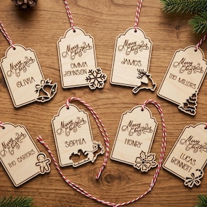Op de afbeelding: Acht lichtgekleurde houten cadeaulabels met rood-witte touwtjes. Elk label is voorzien van de gegraveerde tekst Merry Christmas, een gepersonaliseerde naam of familienaam en een uniek vakantie-icoon zoals een bel, sneeuwvlok of rendier.