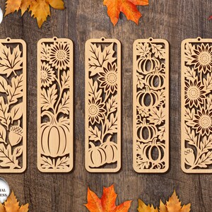 May include: Five wooden bookmarks with intricate cut-out designs. The designs include leaves, sunflowers, pumpkins, and acorns. The bookmarks are light brown and have a small hole at the top. The text "5 DESIGNS" and "ANY MATERIAL THICKNESS" are in the image.
