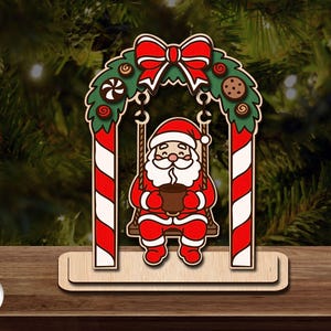 May include: A wooden Christmas decoration featuring Santa Claus on a swing, holding a mug. The swing is framed by red and white striped columns and a green wreath with a red bow. The base has the text "3mm MATERIAL".