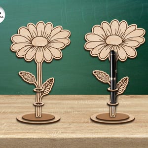 Pu&ograve; includere: Due portapenne in legno a forma di fiore, con uno spessore del materiale di 3 mm. Ogni portapenne presenta un motivo di margherita con una fessura per la penna al centro. I portapenne sono su una superficie di legno.