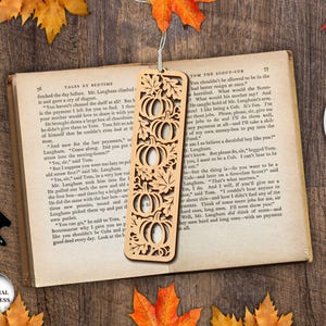 May include: A wooden bookmark with a pumpkin design is placed on an open book. The bookmark has a series of carved pumpkins and leaves. The book is surrounded by fall leaves and a cup of coffee.