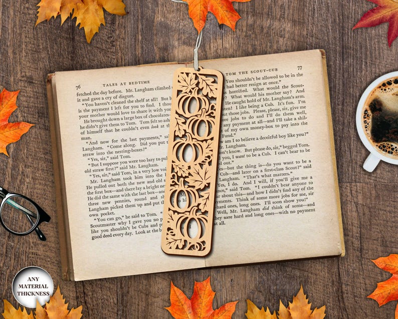 May include: A wooden bookmark with a pumpkin design is placed on an open book. The bookmark has a series of carved pumpkins and leaves. The book is surrounded by fall leaves and a cup of coffee.