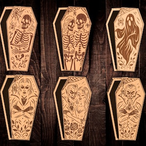 May include: Six wooden coffin-shaped decorations with black interiors, each featuring a different Halloween-themed design. The designs include a skeleton, a pair of skeletons, a ghost, a vampire, a couple, and a vampire. The text "6 Designs" is in the upper left corner.