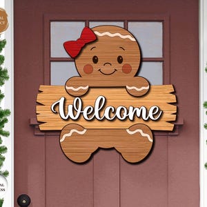 Pode incluir: Um cabide de porta de homem de gengibre de madeira com um laço vermelho e a palavra "Welcome" em escrita branca. O homem de gengibre tem bochechas rosadas e segura a placa. A porta é de cor vermelho escuro.