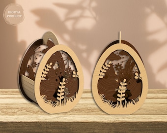 Könnte beinhalten: Zwei hölzerne Ostereier-förmige Boxen mit filigranen Ausschnitten von Hasen, Blumen und Blättern. Die Boxen sind braun und haben eine natürliche Holzoptik.