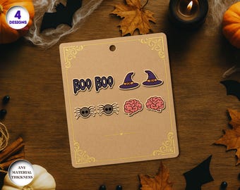 May include: A Halloween-themed display featuring four earring designs: "BOO BOO" text, witch hats, spiders, and brains. The earrings are displayed on a brown card with gold decorative corners, set against a wooden background with pumpkins and leaves.