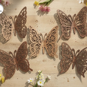 Puede incluir: Cinco adornos de madera con forma de mariposa, con diseños intrincados. Las mariposas, en tonos marrones, presentan patrones florales y geométricos. Se exhiben sobre una superficie de madera clara, rodeadas de flores.