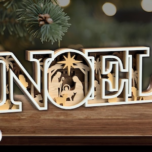 May include: A layered wooden Christmas decoration spelling "NOEL". The letter "O" contains a nativity scene with a star. The letters have white edges and natural wood interiors, set on a wooden surface, with a Christmas tree in the background.