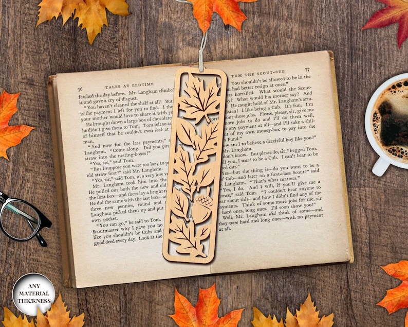 May include: A wooden bookmark with a leaf and acorn design, resting on an open book. The bookmark is light brown and has cut-out details. The book is open to pages with text. Autumn leaves and a cup of coffee complete the scene.