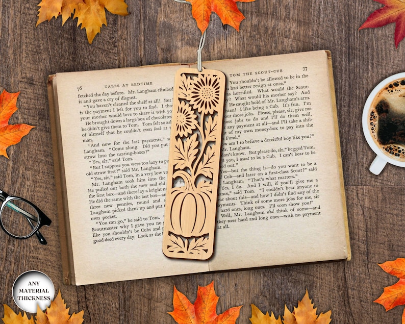 May include: A wooden bookmark with a laser-cut design featuring sunflowers, leaves, and a pumpkin. The bookmark is placed on an open book with autumn leaves and a cup of coffee in the background.