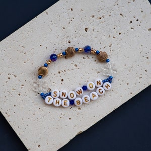 May include: A white beaded bracelet with gold lettering spelling "SNOW ON THE BEACH" and blue and gold beads.