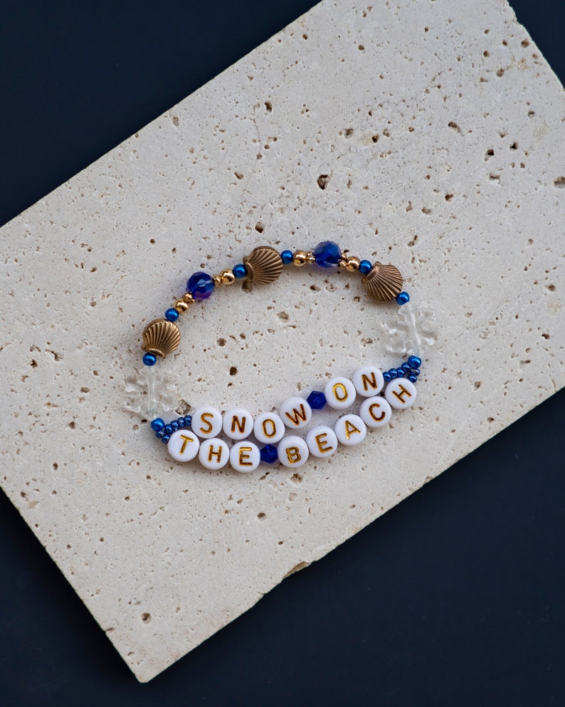 May include: A white beaded bracelet with gold lettering spelling "SNOW ON THE BEACH" and blue and gold beads.