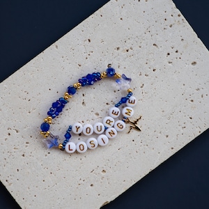 May include: A blue and gold beaded bracelet with white beads spelling out "You're Losing Me" and a gold airplane charm.