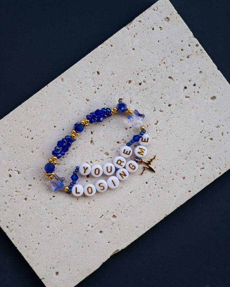 May include: A blue and gold beaded bracelet with white beads spelling out "You're Losing Me" and a gold airplane charm.