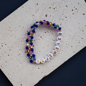 May include: A multi-colored beaded bracelet with white beads that spell out "BE JEWELED".