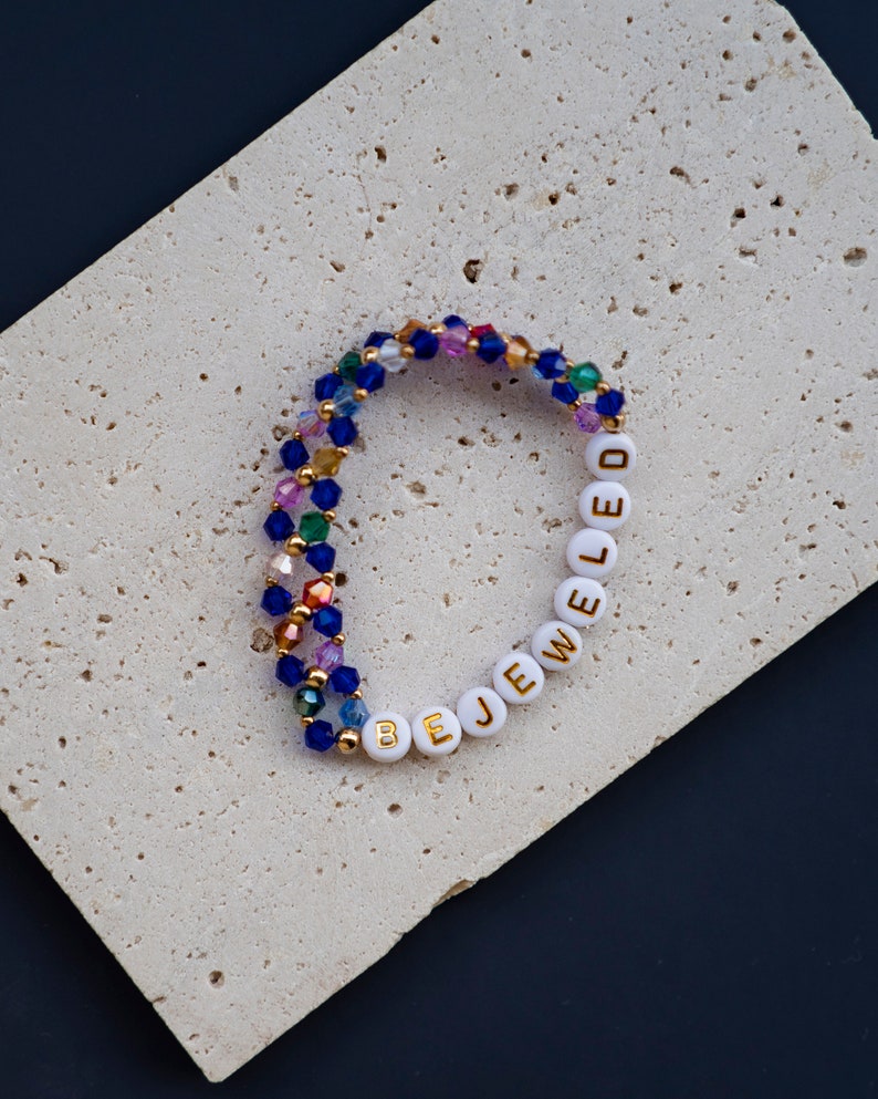 May include: A multi-colored beaded bracelet with white beads that spell out "BE JEWELED".