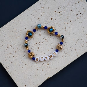 May include: A gold beaded bracelet with blue and gold beads and a charm with the letters "BTTWS" in gold lettering.