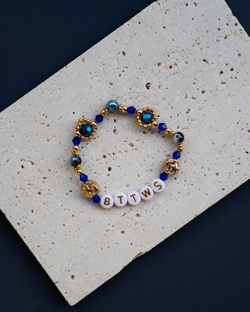 May include: A gold beaded bracelet with blue and gold beads and a charm with the letters "BTTWS" in gold lettering.