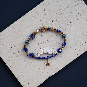 May include: A blue and gold beaded bracelet with a charm of the Eiffel Tower and white beads spelling out "PARIS".